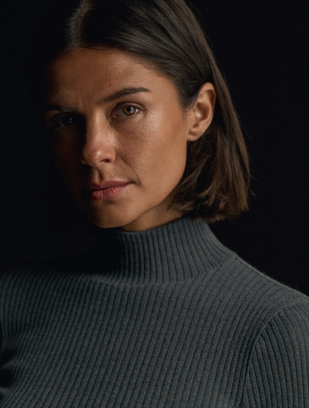 Editorial portrait of the composed persona — a woman in a charcoal turtleneck, half-lit against a near-black backdrop